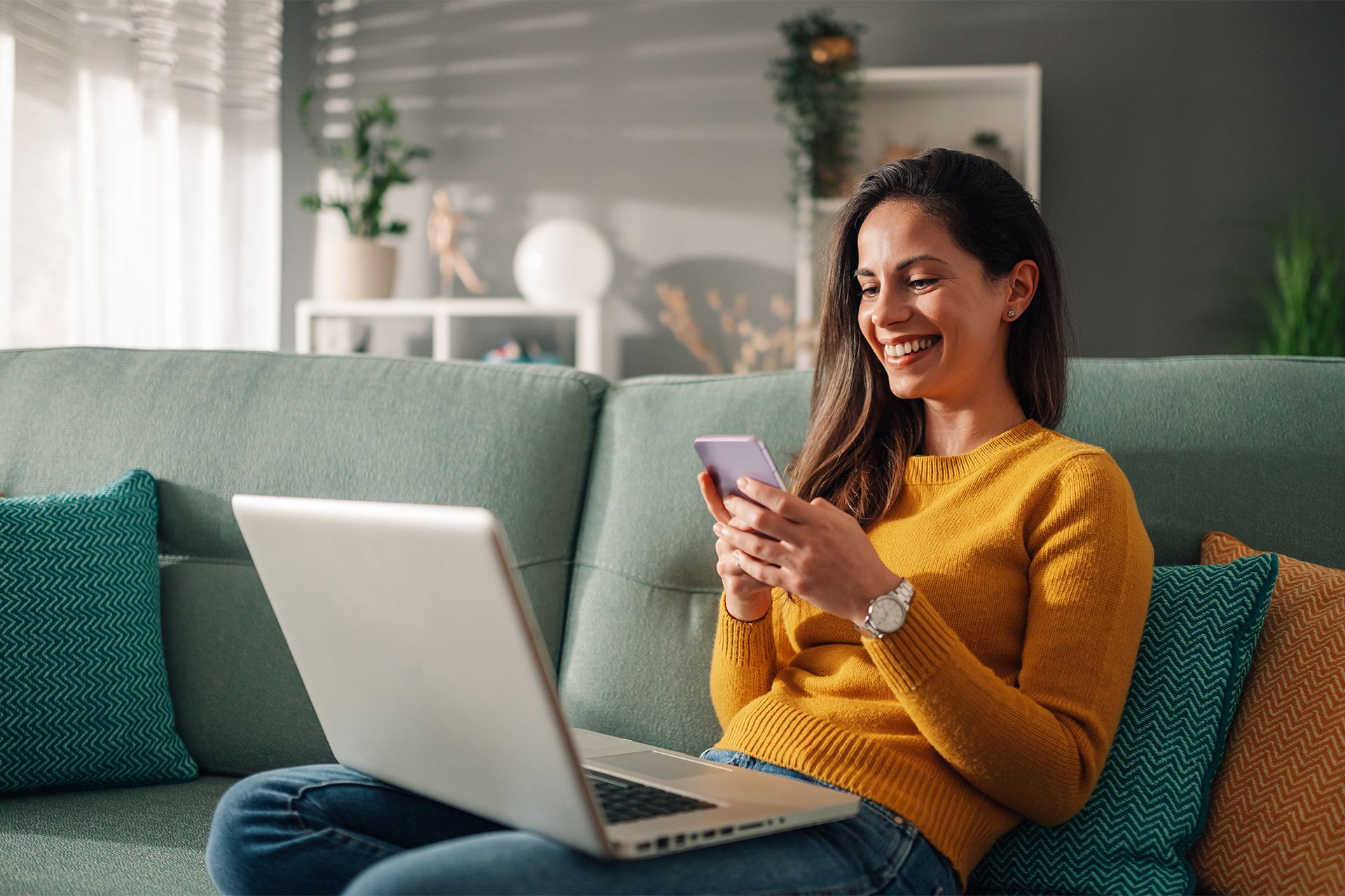 Woman sitting on her couch at home with her laptop and phone, smiling