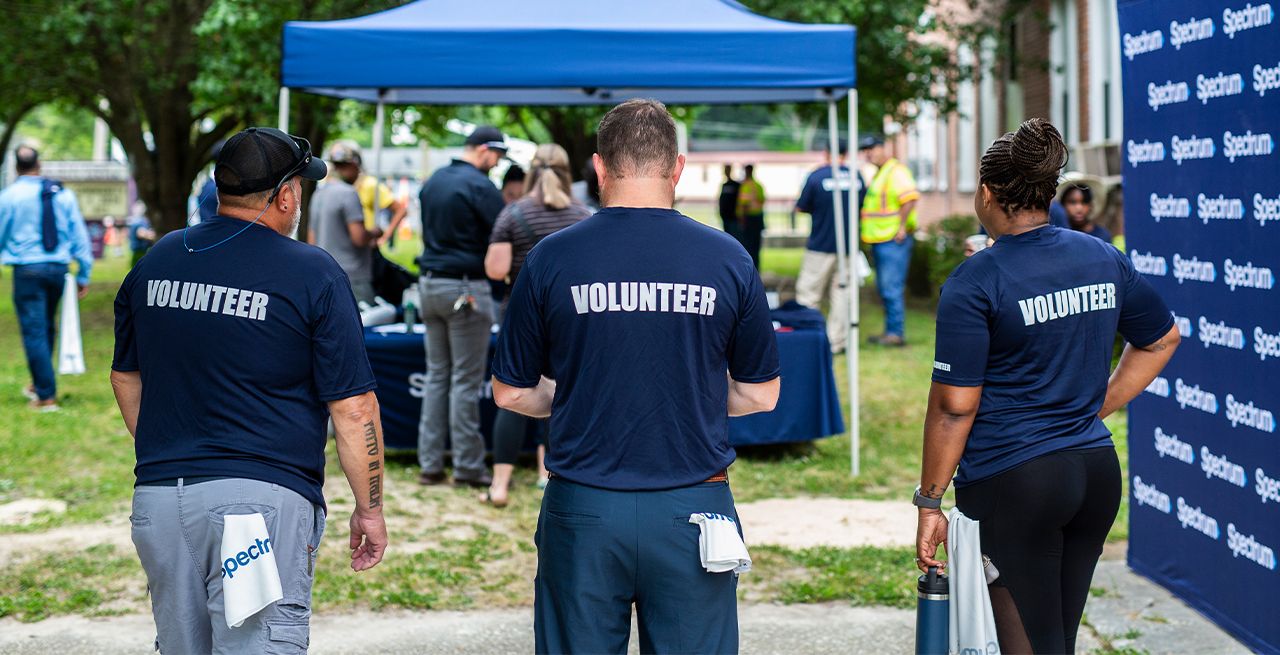 Spectrum volunteers on the grounds of a community center, standing in a row, backs turned, with "Volunteer" t-shirts on